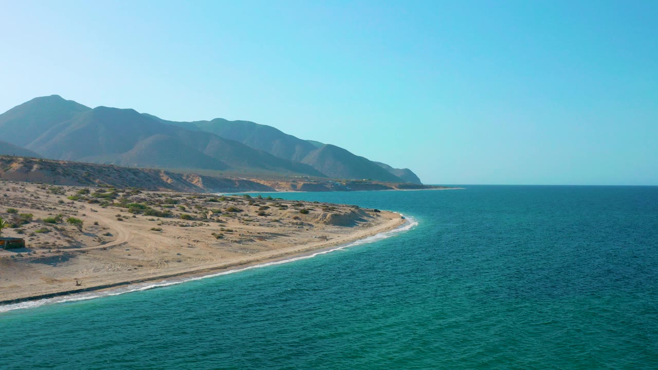 Opening aerial view over El Sargento La Ventana with blue ocean on the right, a curving sandy shoreline on the left, distant mountains, clear sky, and no visible people.