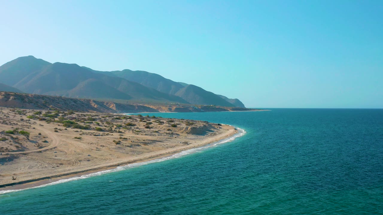 Mid-clip aerial frame over El Sargento La Ventana showing scrub-covered sand in the foreground, blue water along the curved shore, hazy mountains, and no visible people.