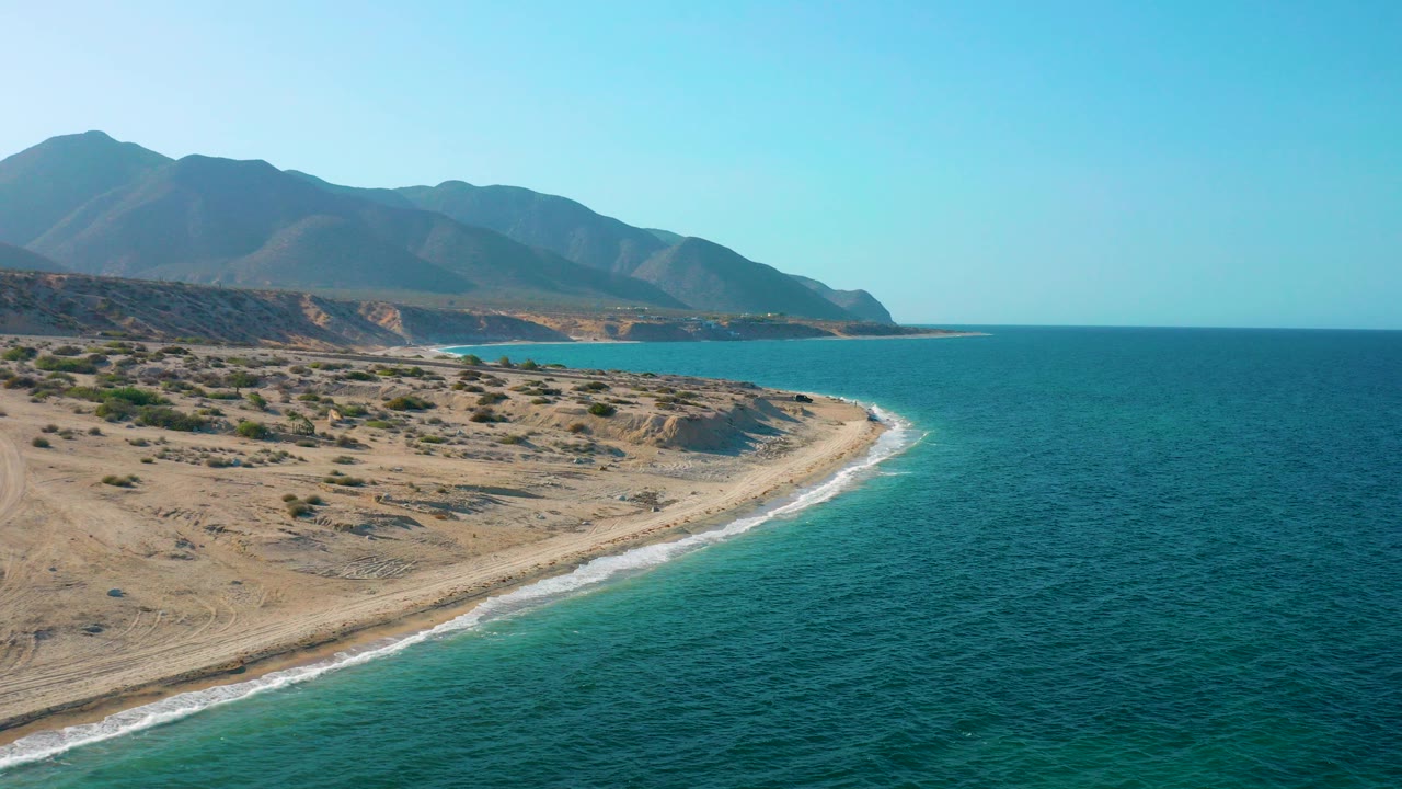 Opening aerial frame over El Sargento La Ventana, Mexico, showing a curved sandy coast, blue green water, low desert brush, distant mountains, and no visible people.