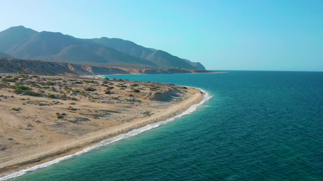 Aerial frame over El Sargento La Ventana, Mexico, showing dry brushland meeting a pale beach, small surf, wide blue ocean, layered mountains, and no visible people.