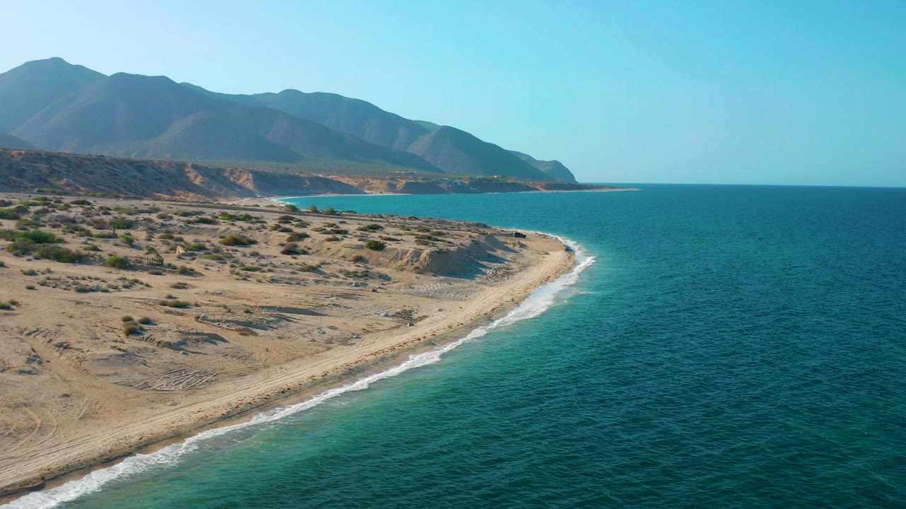 Mid-clip aerial frame over El Sargento La Ventana, Mexico, with mountains occupying the left background, a curved beach below, blue ocean on the right, and no visible people.