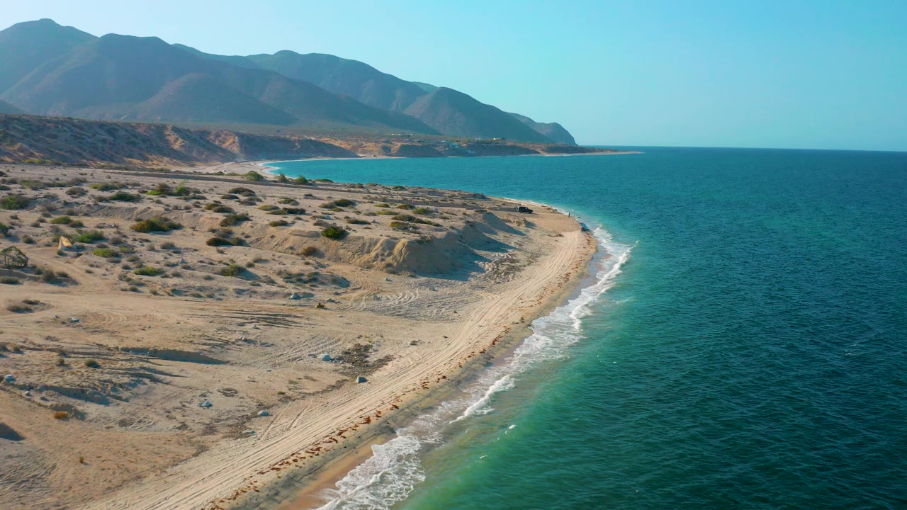 Aerial view over El Sargento La Ventana, Mexico, showing a pale sandy beach curving along blue-green water, desert scrub, distant mountains, and no visible people.