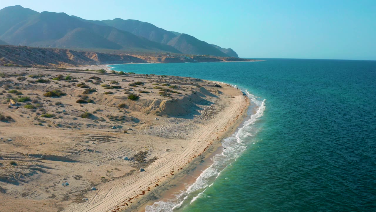 Drone perspective over El Sargento La Ventana, Mexico, showing empty sand with vehicle tracks, small white waves on the right, open sea, sparse shrubs, distant headland, and no visible people.