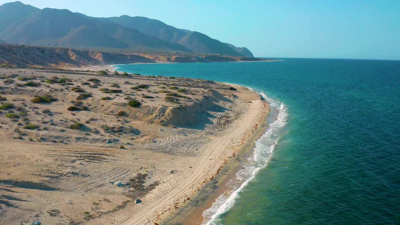 Closer aerial view of El Sargento La Ventana, Mexico, with pale dunes and scrub in the foreground, a curved beach beside blue water, mountains across the background, and no visible people.