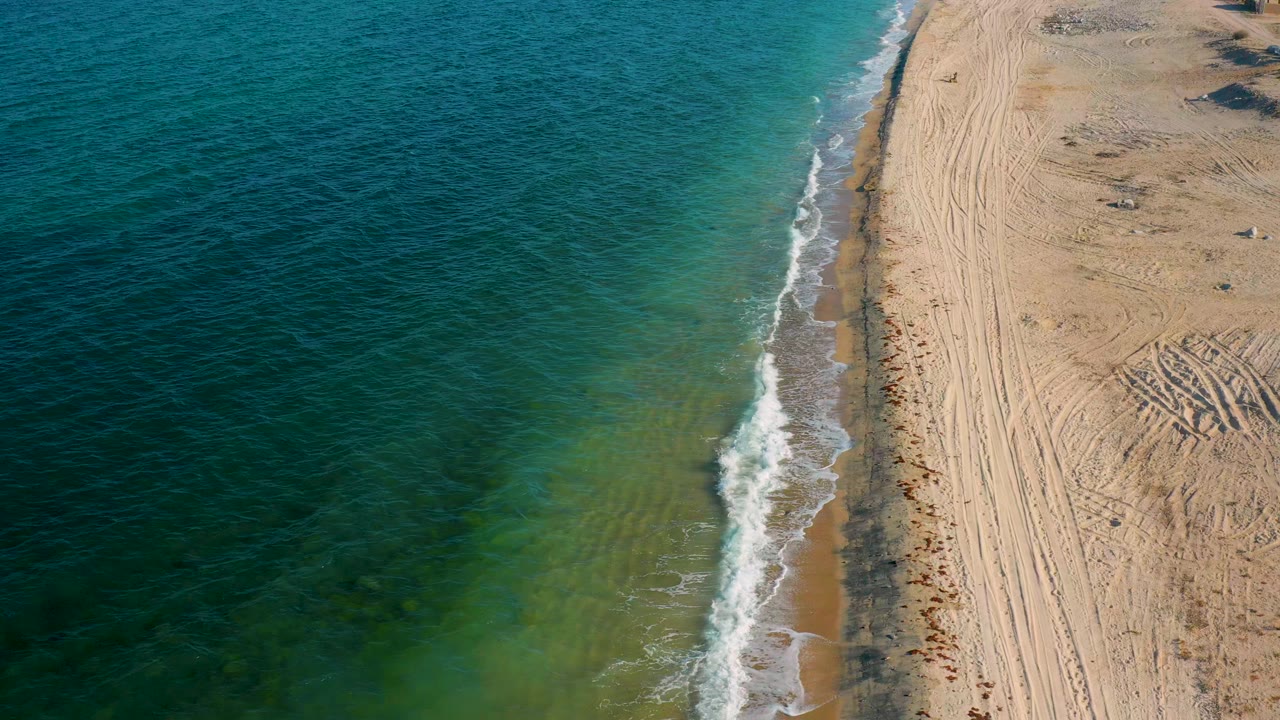 Aerial frame from El Sargento La Ventana showing a foamy wave advancing along the narrow shoreline beside tire-marked sand, with no visible people.