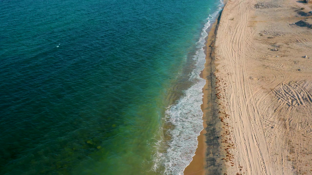 Overhead frame of El Sargento La Ventana with a larger white surf patch rolling toward the beach, textured water to the left, and no visible people.