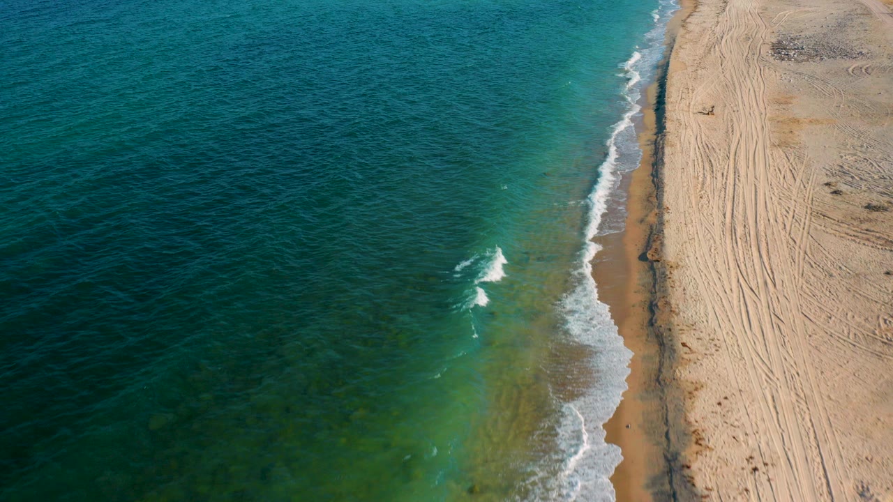 Elevated view over El Sargento La Ventana, Mexico, with green-blue water on the left, pale sand on the right, a thin surf line, and no visible people.