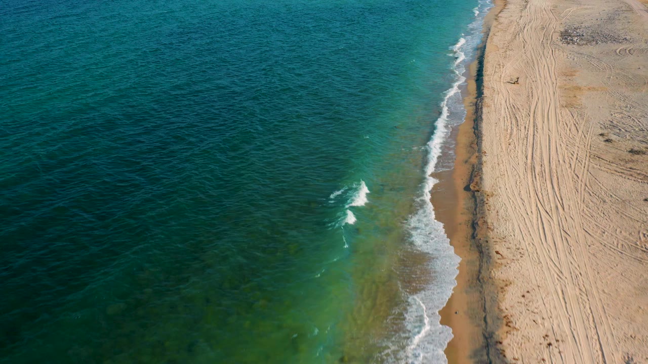 Aerial frame over El Sargento La Ventana, Mexico, showing a small white breaker in green water beside a narrow sandy beach, with no visible people.