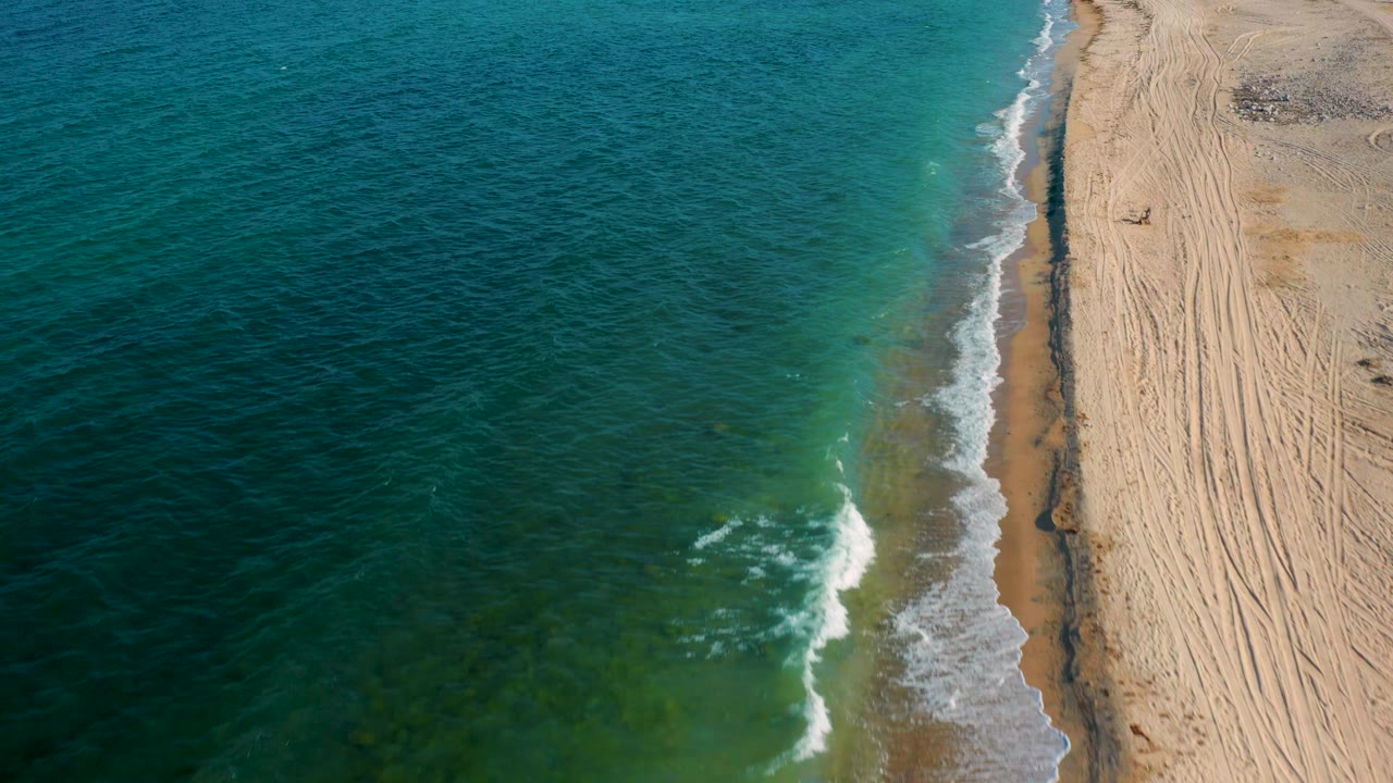 Overhead coastal view in El Sargento La Ventana, Mexico, with teal shallows, darker ocean water, a curving foam edge, tire-marked sand, and no visible people.