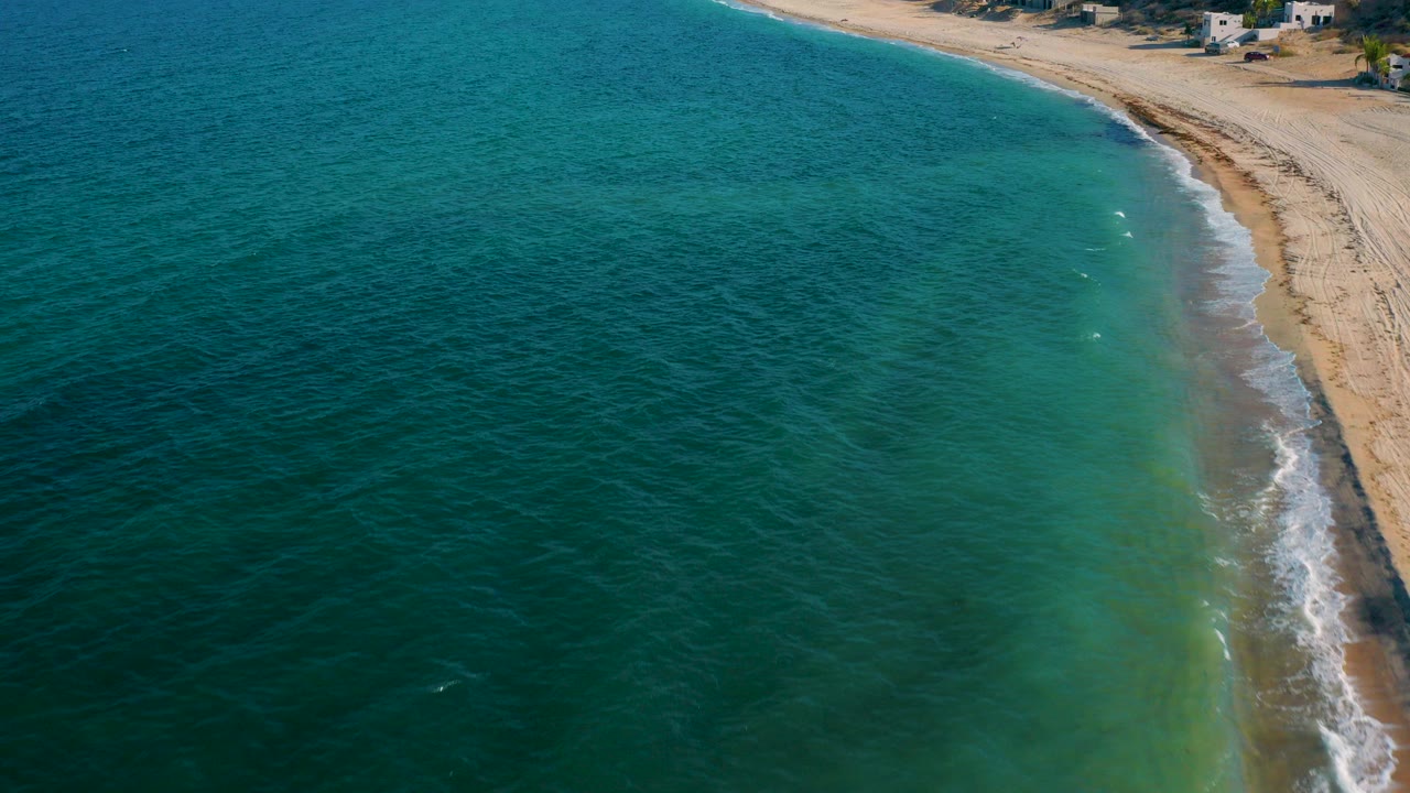 Opening aerial frame over El Sargento La Ventana, Mexico, with blue-green water filling the left side, a pale sandy curve at right, hillside homes near the top edge, and no visible people.