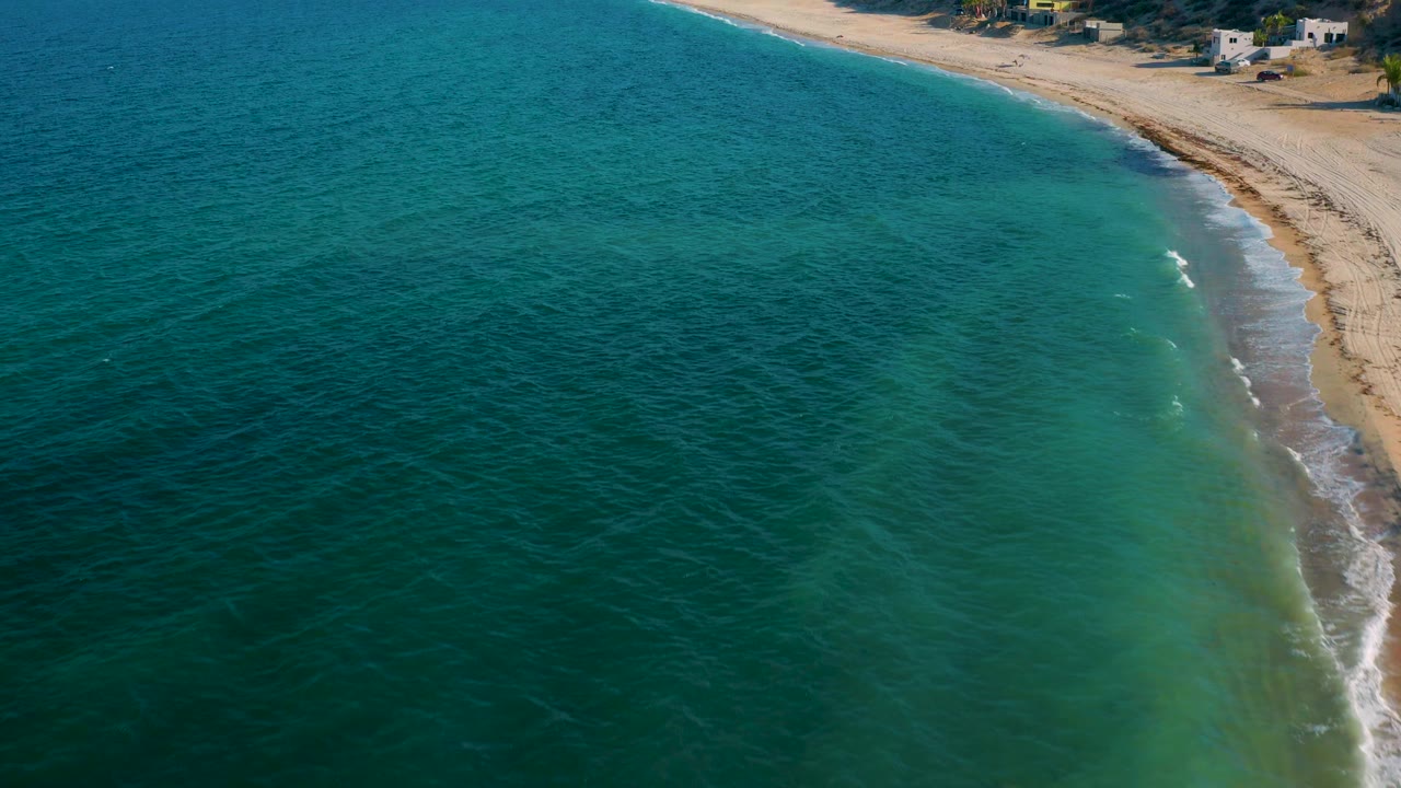 Frame over El Sargento La Ventana, Mexico, showing darker blue water offshore, greener shallows near the sand, a thin line of foam on the curved beach, small homes above the shore, and no visible people.