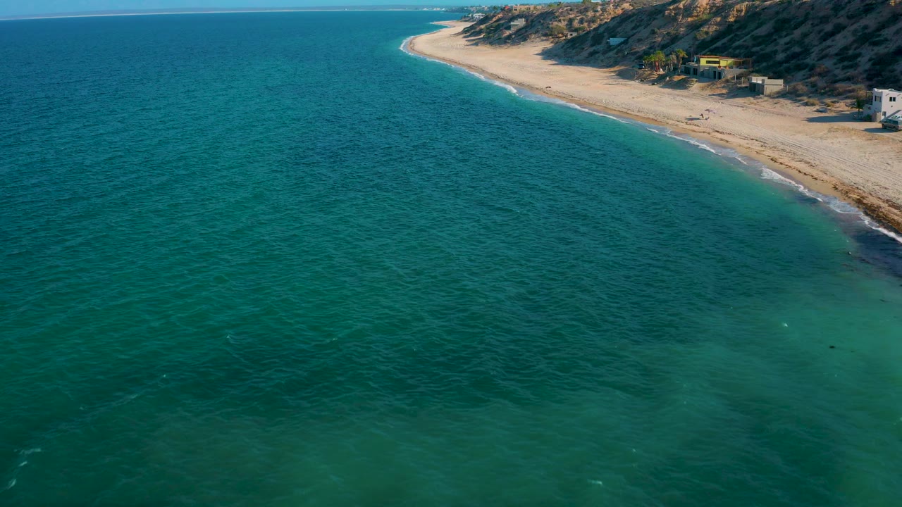 Turquoise sea and pale beach curve below a rocky slope with scattered homes.