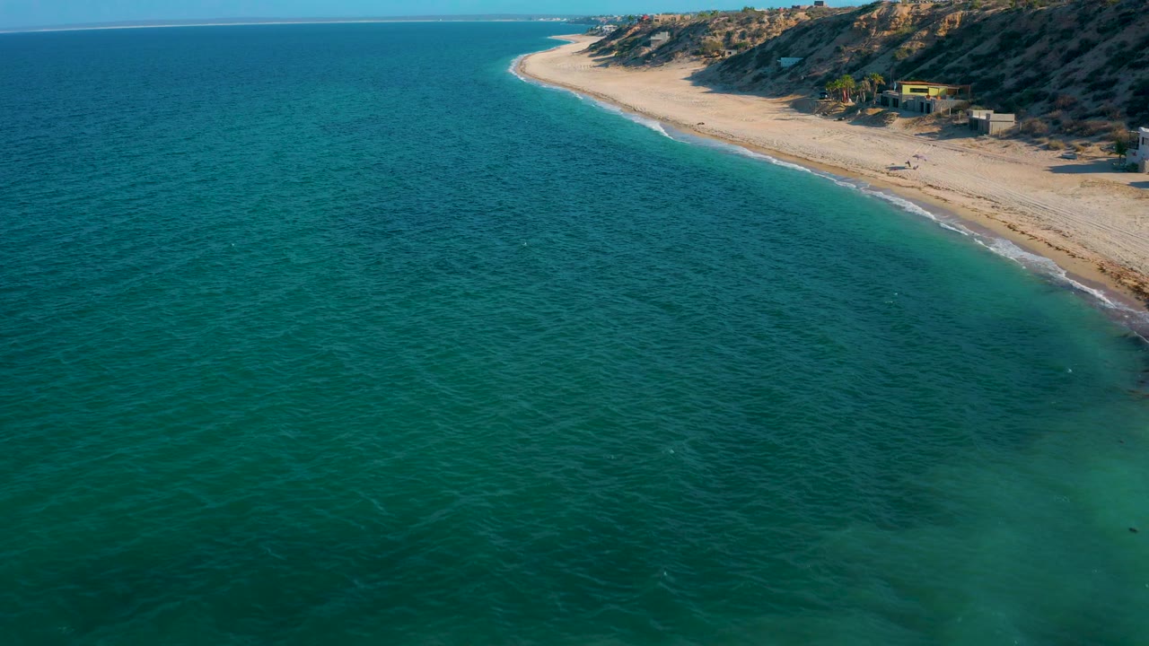 Wide aerial view over El Sargento La Ventana with textured ocean water across the foreground, a sandy beach along the right edge, bluffside homes, and no visible people.