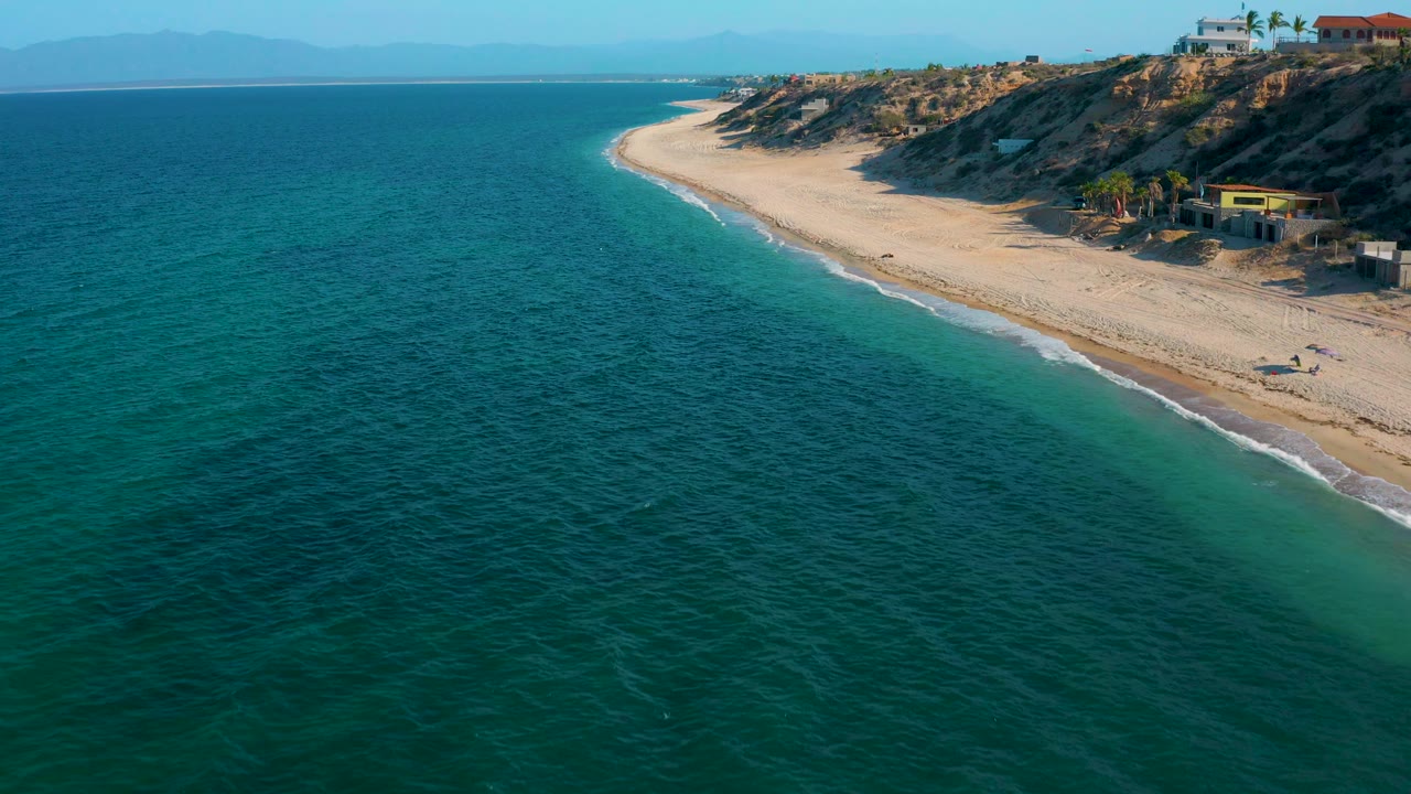 Elevated coastal frame from El Sargento La Ventana, Mexico, showing textured blue-green water, a narrow white surf edge, pale sand, blufftop buildings, distant mountains, and no visible people.