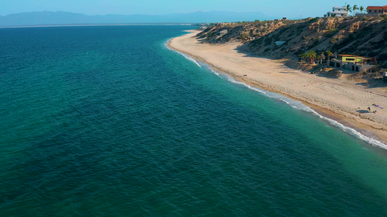 Drone-style view over El Sargento La Ventana, Mexico, with open turquoise water covering most of the frame, a sandy beach along the right, rocky slopes above it, small coastal structures, and no visible people.