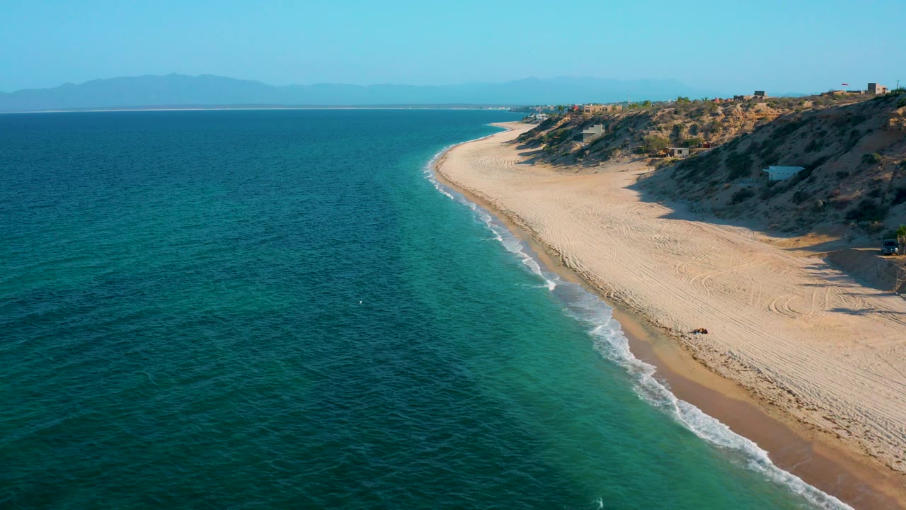 An aerial coastline frame shows open blue water dominating the left foreground, pale sand on the right, a narrow foam line, low hillside scrub, and no visible people.