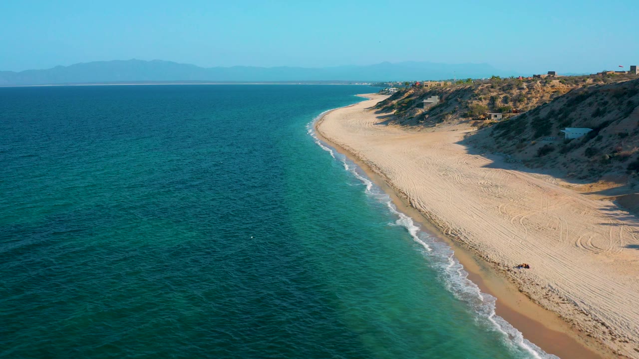 A high coastal view in El Sargento La Ventana, Mexico shows empty sand, dark blue sea, a soft turquoise band near shore, dune shadows, and no visible people.