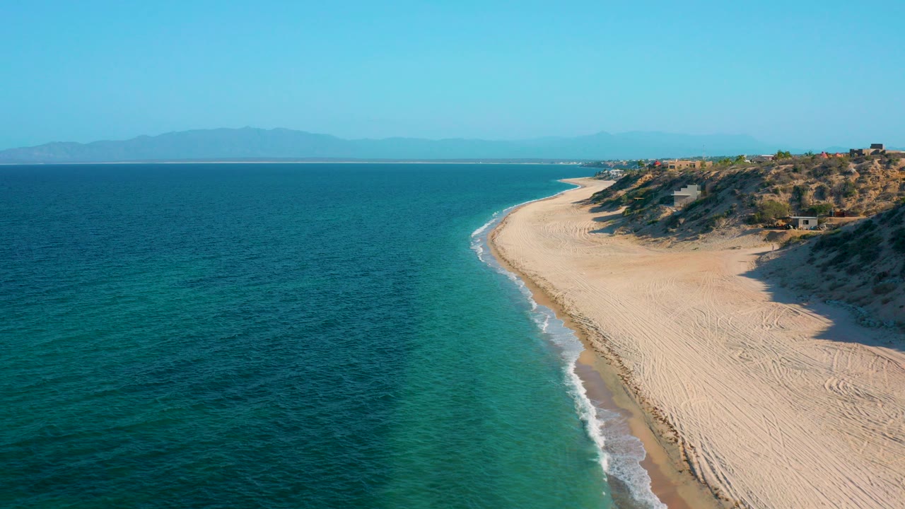 Opening aerial frame over El Sargento La Ventana, Mexico, with turquoise water on the left, a curving pale beach on the right, distant mountains on the horizon, and no visible people.