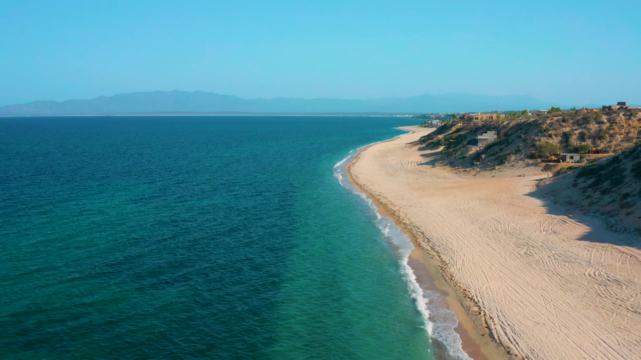 Aerial frame in El Sargento La Ventana, Mexico, showing the shoreline curve more centrally, with textured sand tracks, low dunes, small blufftop buildings, and no visible people.