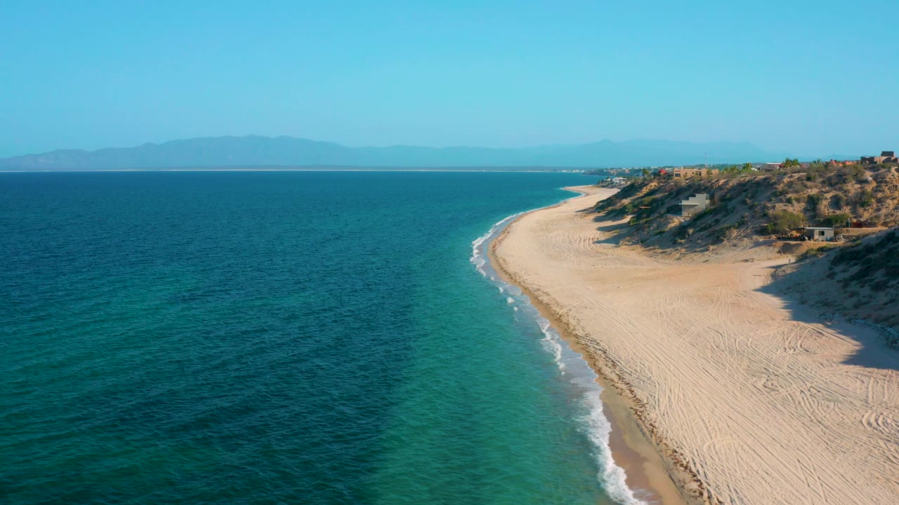 High aerial view over El Sargento La Ventana, Mexico, with deep blue water across the left, a narrow foamy surf edge, dunes and sparse vegetation to the right, and no visible people.