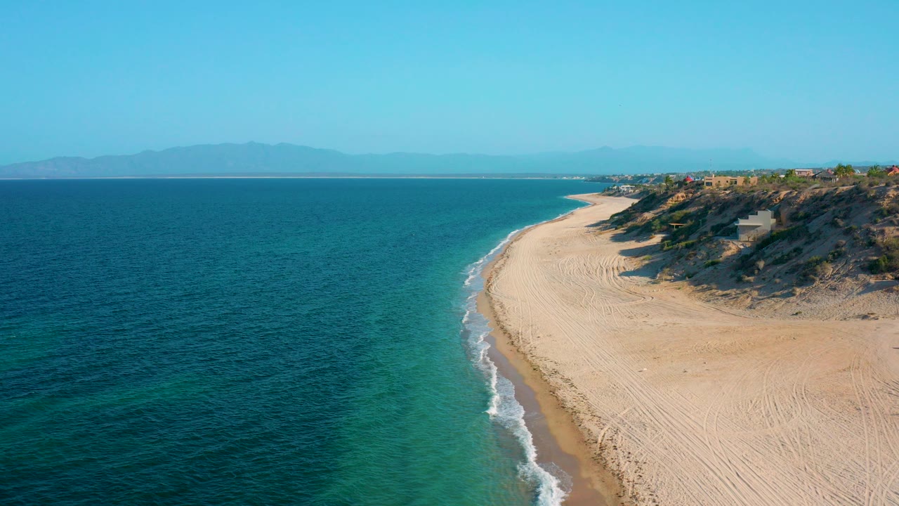Aerial frame from El Sargento La Ventana, Mexico, showing blue green ocean on the left, a pale curved beach on the right, low cliffs beyond the sand, distant mountains, and no visible people.