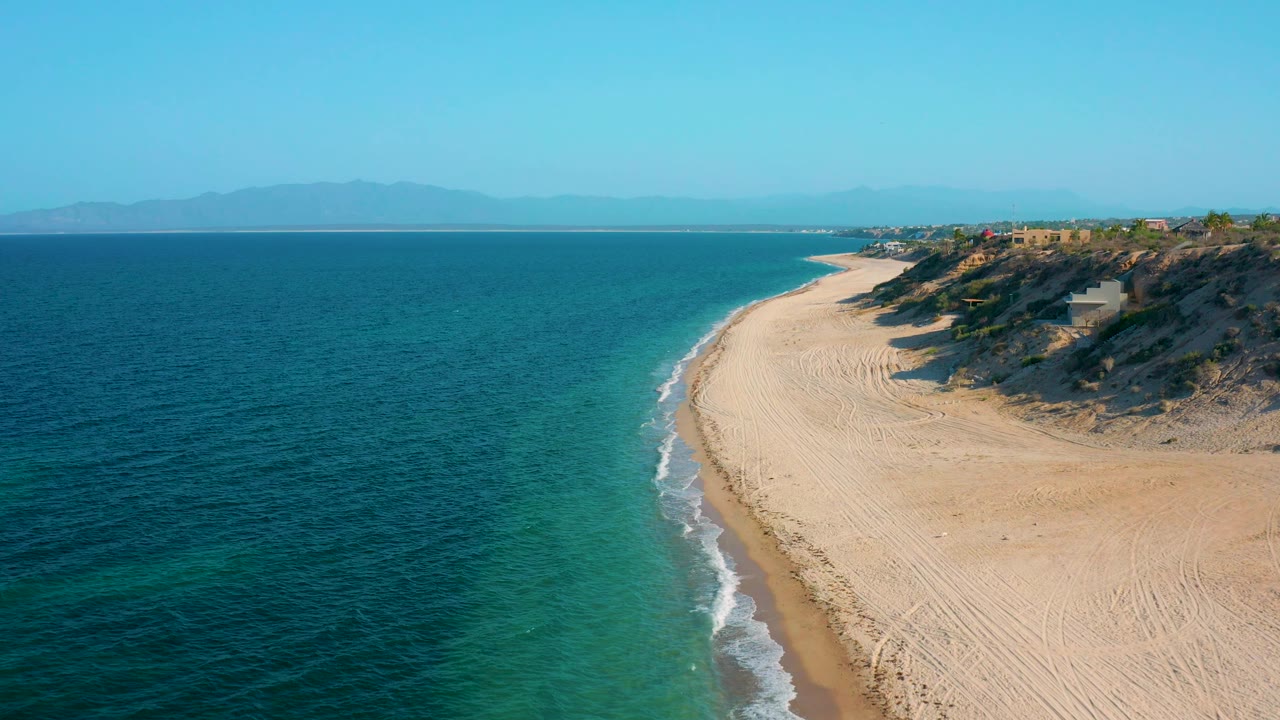 Aerial frame from El Sargento La Ventana, Mexico, where a thin white surf line follows the beach, the ocean opens across the left side, blufftop buildings sit above the sand, and no visible people appear.