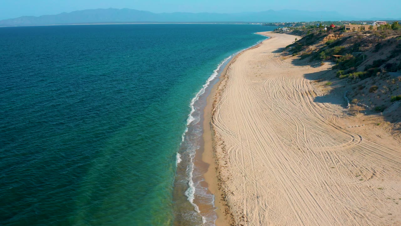 El Sargento La Ventana, Mexico aerial frame showing a curving beach, darker offshore water, light aqua shallows, white shorebreak, bluff-top buildings, and no visible people.