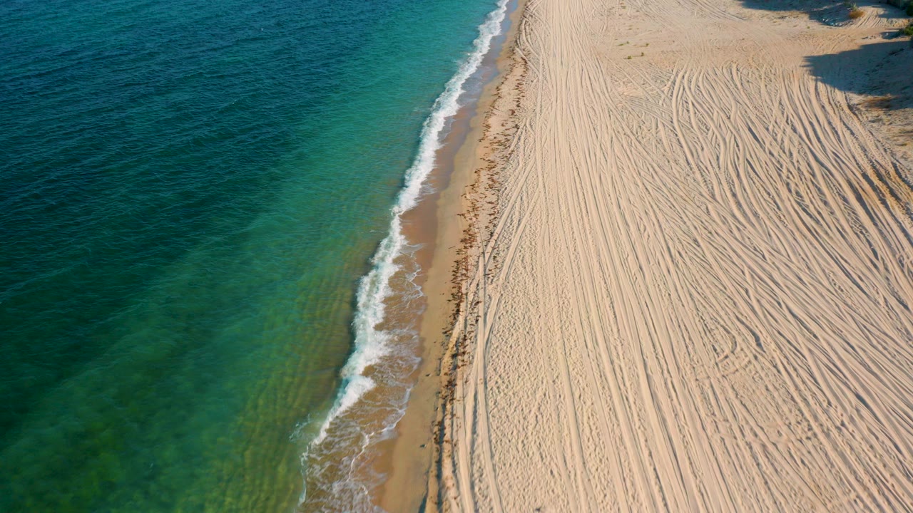 High aerial view in El Sargento La Ventana, Mexico, showing green-blue ocean on the left, pale tracked sand on the right, and no visible people.