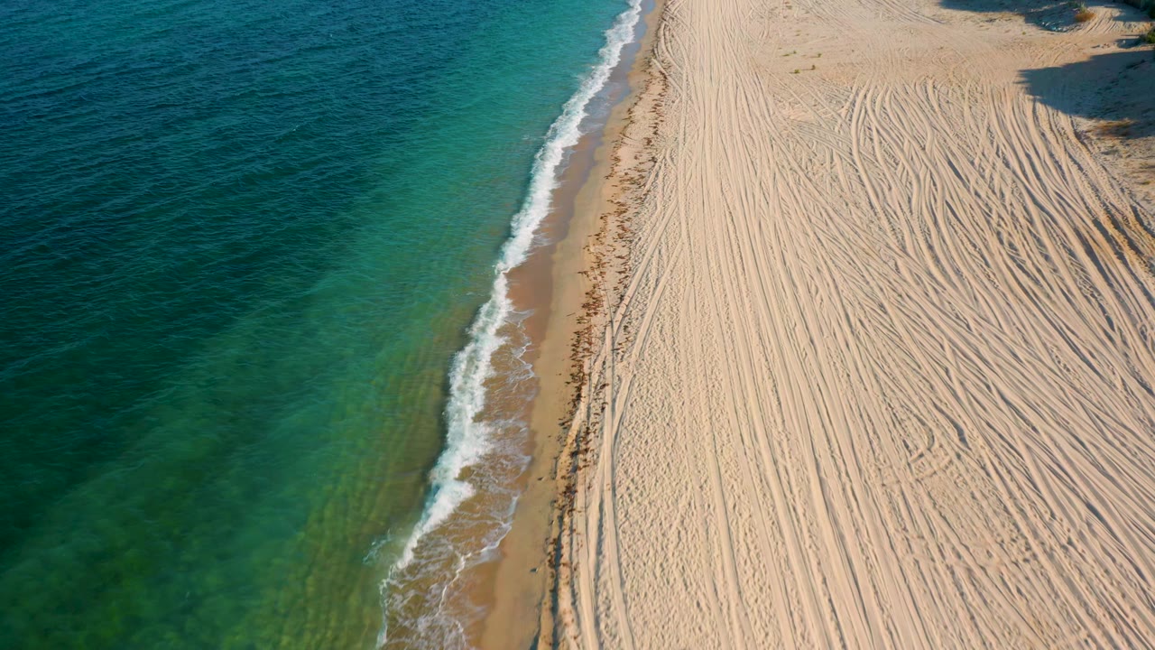 Aerial shoreline frame from El Sargento La Ventana, Mexico, with shallow green water, a thin foam edge, broad pale sand, and no visible people.