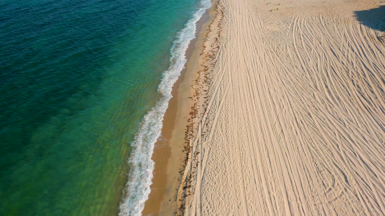 Overhead drone frame in El Sargento La Ventana, Mexico, where the surf line curves along clear water and heavily marked sand, with no visible people.