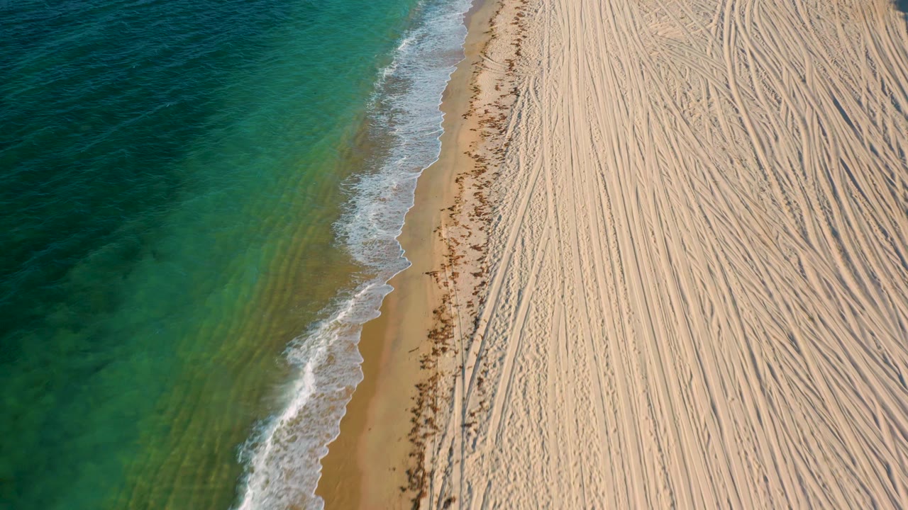Aerial coastline view with turquoise water, foam, wet sand, and tire-marked beach.