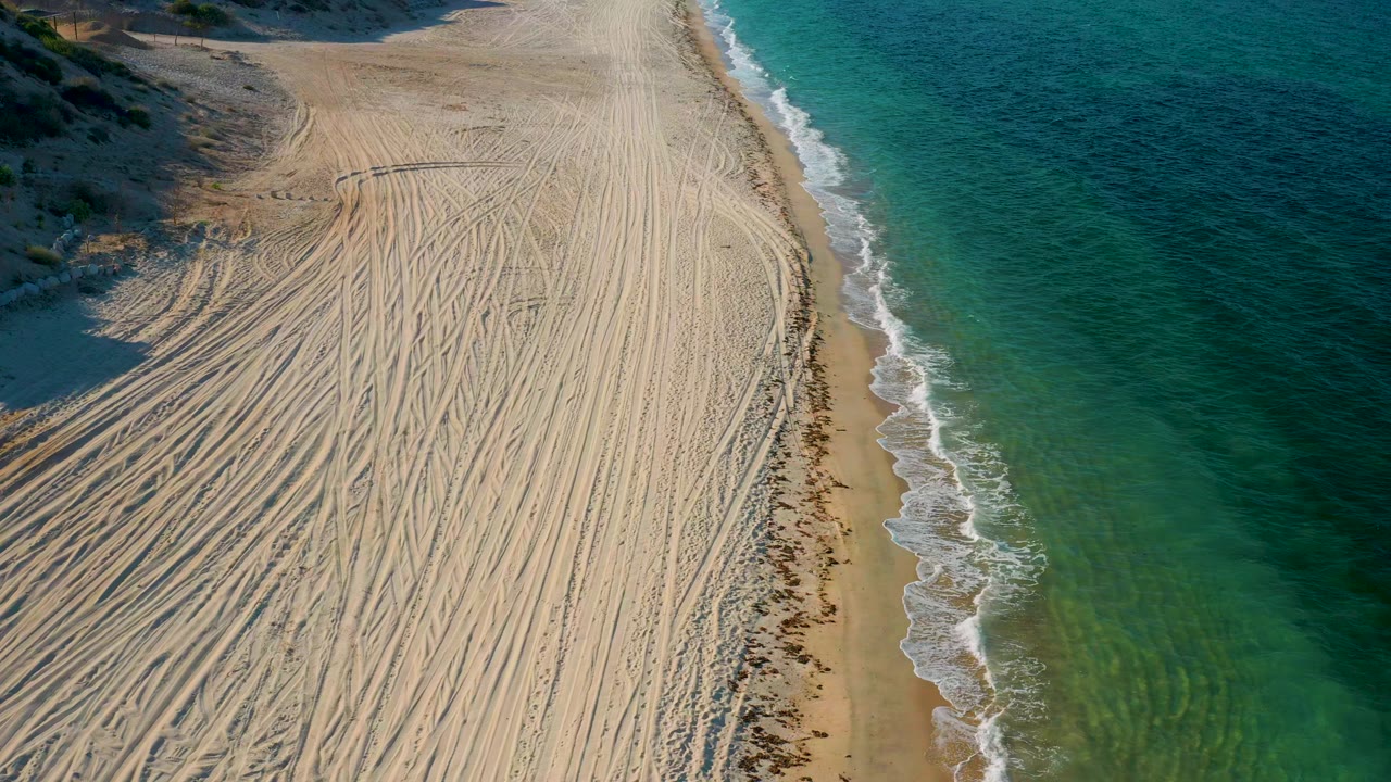 The aerial frame holds over the Mexico shoreline with dense track marks across the sand, small white waves along the edge, clear green-blue water, and no visible people.
