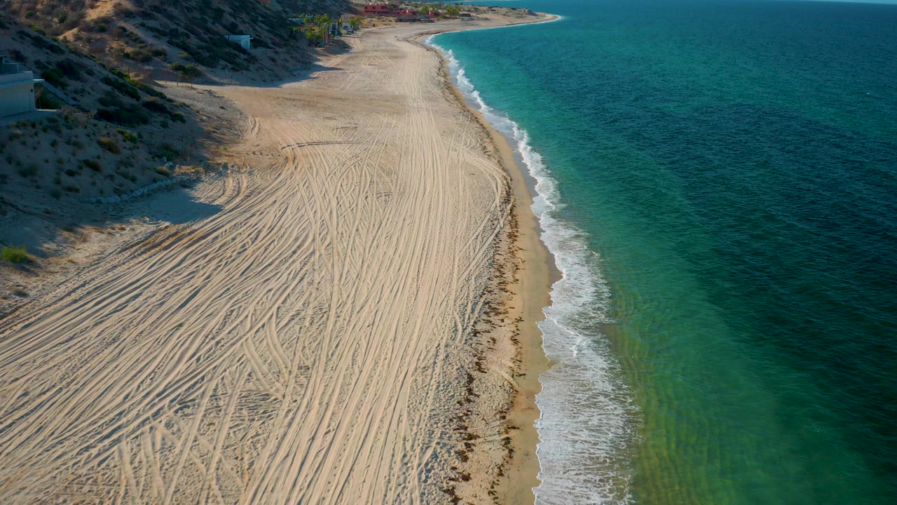 Aerial view over El Sargento La Ventana, Mexico, showing a tire-tracked sandy beach below, blue-green water on the right, hillside terrain on the left, and no visible people.