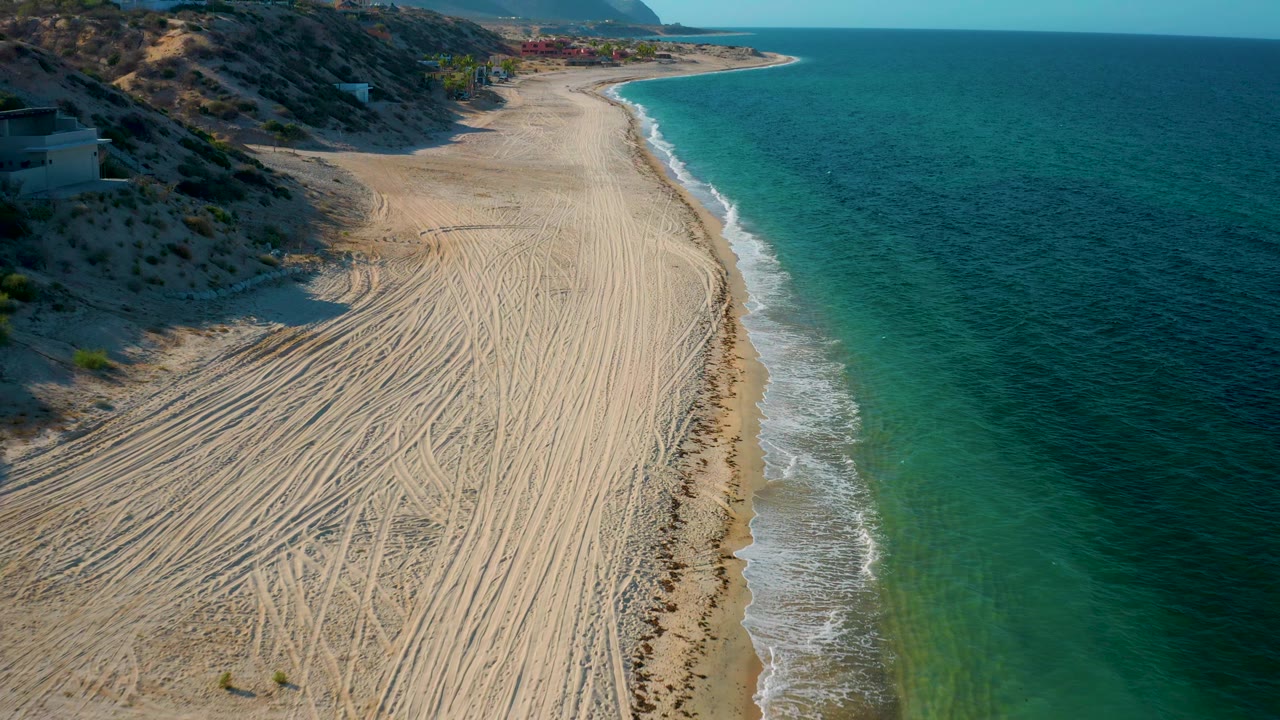 Drone view along the curving beach at El Sargento La Ventana, Mexico, with clear water to the right, tracked sand below, low hills to the left, and no visible people.