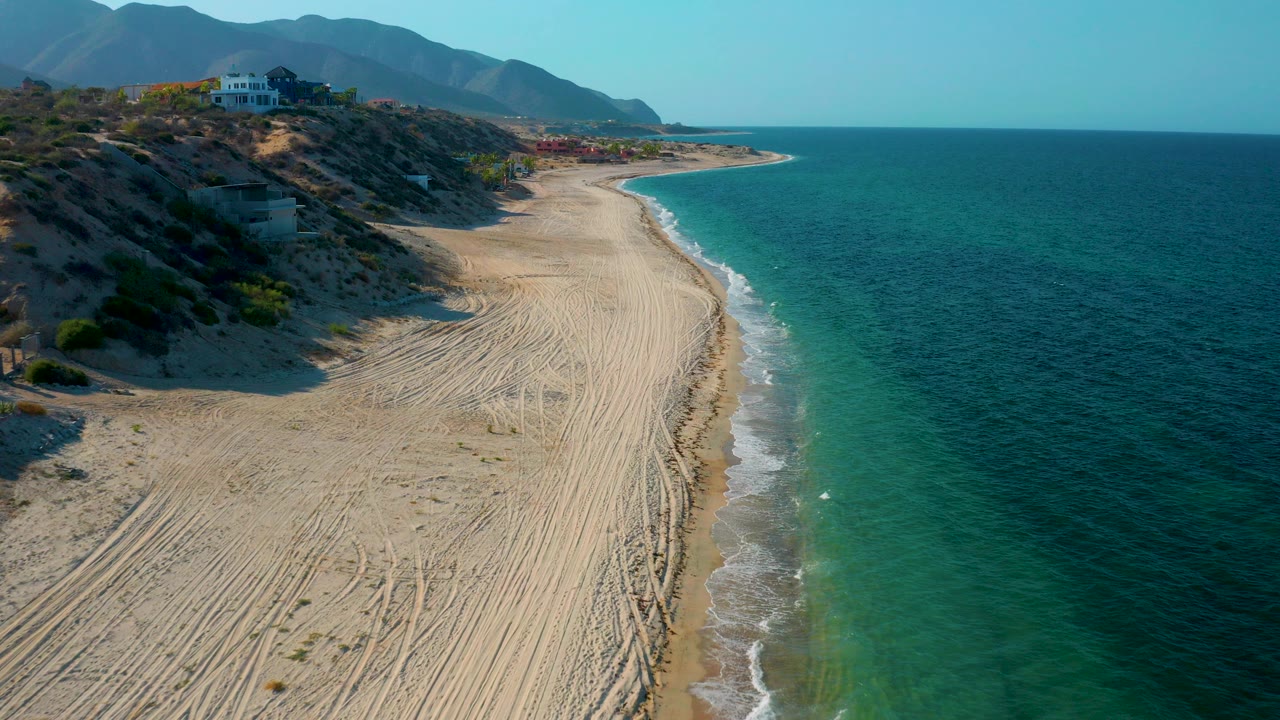 Opening aerial frame over El Sargento La Ventana, Mexico, showing a wide sandy beach with tire tracks, turquoise surf, scrubby dunes, hillside buildings, distant mountains, and no visible people.