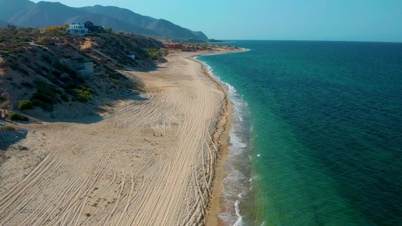 One second into the clip, the aerial view holds on the El Sargento La Ventana shoreline with patterned sand tracks, a narrow surf edge, blue-green water, hillside buildings, mountains, and no visible people.