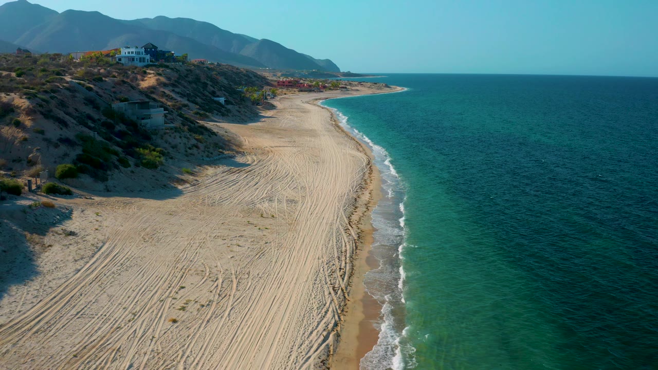 At 0:02, the frame shows the curving beach in El Sargento La Ventana, Mexico, with clear water to the right, dry dunes and hillside buildings to the left, mountains behind, and no visible people.