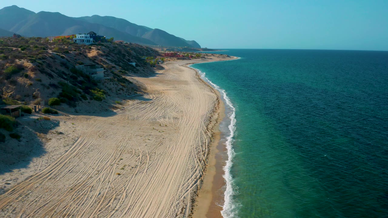 Aerial coastline with beach tracks, gentle surf, blue ocean, hillside development, and mountains.