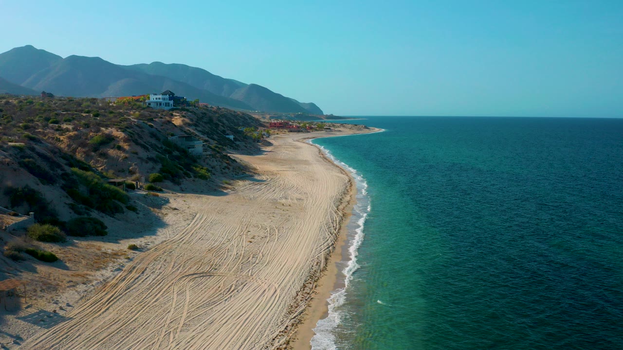 Opening aerial frame over El Sargento La Ventana, Mexico, with a wide sandy beach curving beside blue ocean, scrub hills, distant mountains, and no visible people.