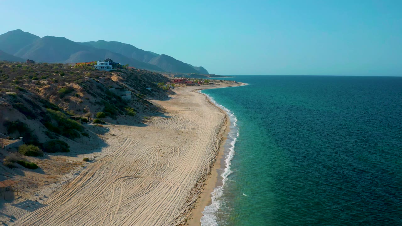 Aerial view in El Sargento La Ventana, Mexico, showing the curved beach edge, pale surf line, turquoise shallows, hillside scrub, distant mountains, and no visible people.