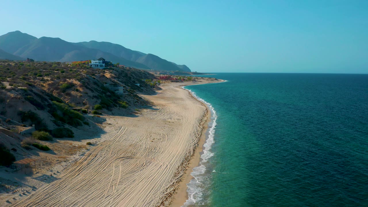 Aerial frame from El Sargento La Ventana, Mexico, with broad sand in the foreground, blue ocean to the right, low surf along the beach, mountain ridges behind the coast, and no visible people.