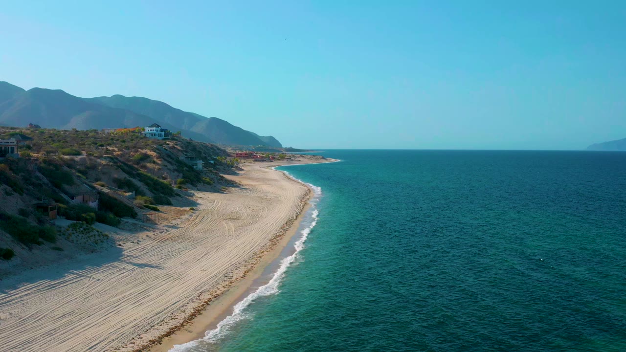 Opening aerial frame over El Sargento La Ventana, Mexico, showing pale sand dunes along the left, blue-green sea on the right, distant mountains, and no visible people.