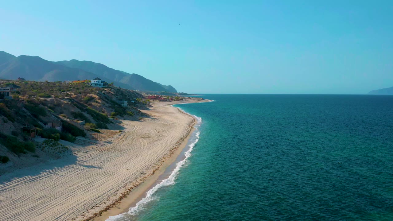 A wide aerial frame in El Sargento La Ventana, Mexico, with the beach curving toward the horizon, turquoise shallows near shore, deeper blue water beyond, and no visible people.