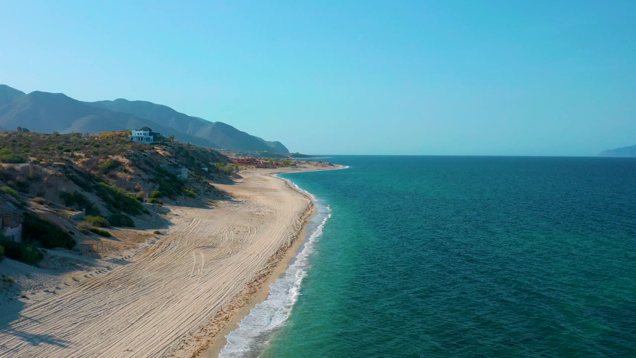 Opening aerial view over El Sargento La Ventana, Mexico, with a wide sandy shoreline, turquoise water at the surf line, darker ocean to the right, mountains in the background, and no visible people.