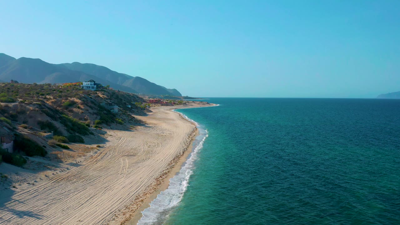 Aerial frame from El Sargento La Ventana, Mexico, showing the beach running diagonally through the frame, foamy water edging the sand, scrub-covered hills at left, distant mountains, and no visible people.