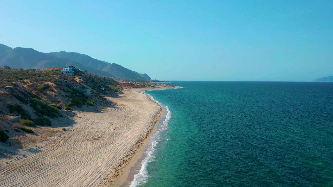 High coastal view of pale sand, breaking waves, ocean, hills, and distant mountains.