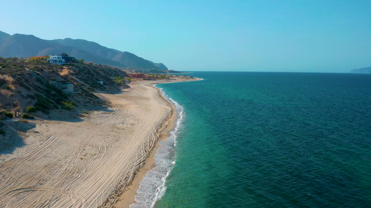 Aerial view over El Sargento La Ventana, Mexico, with pale tracked sand, a thin white surf edge, turquoise shallows, dark blue water, hillside buildings, mountains, and no visible people.