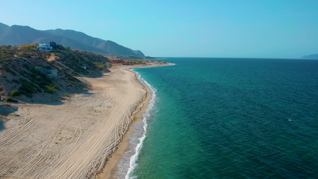 Drone-like aerial frame over El Sargento La Ventana, Mexico, with the shoreline running diagonally, hillside homes above the sand, distant mountains, turquoise coastal water, and no visible people.