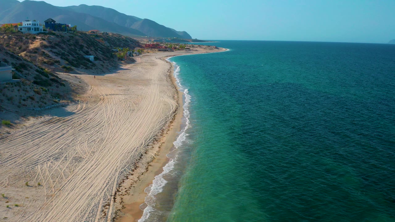 Opening aerial frame over El Sargento La Ventana, Mexico, with raked sand in the foreground, turquoise water to the right, blufftop buildings to the left, distant mountains, and no visible people.