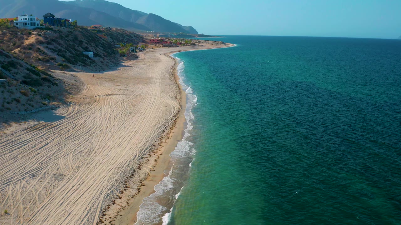 Aerial frame over El Sargento La Ventana, Mexico, with the beach running diagonally through the frame, green-blue shallows beside deeper water, dry bluffs with buildings, mountains beyond, and no visible people.